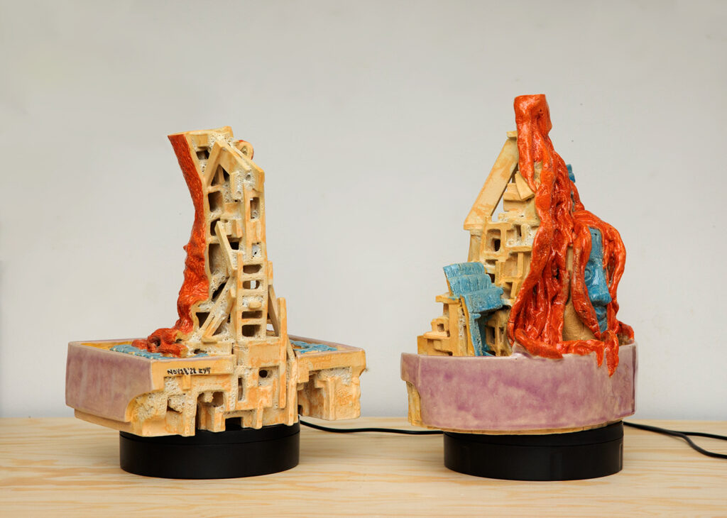 Two red, blue, and lavender glazed ceramic pieces on turntables, turned to reveal front and rear three-quarters' views. One features a tree root system partially covering the carving of a face, the other reveals a scaffolding-like structure on the back of a tree trunk growing out of a tiled sidewalk.